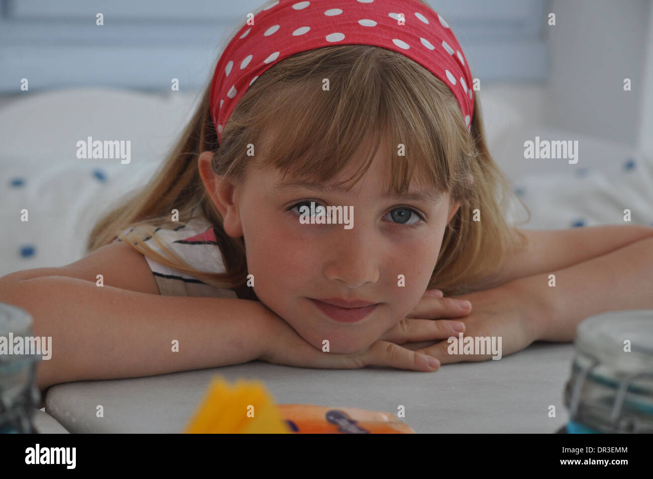 portrait of pretty little blonde girl resting her head on her hands while wearing a red polka dot hair band Stock Photo