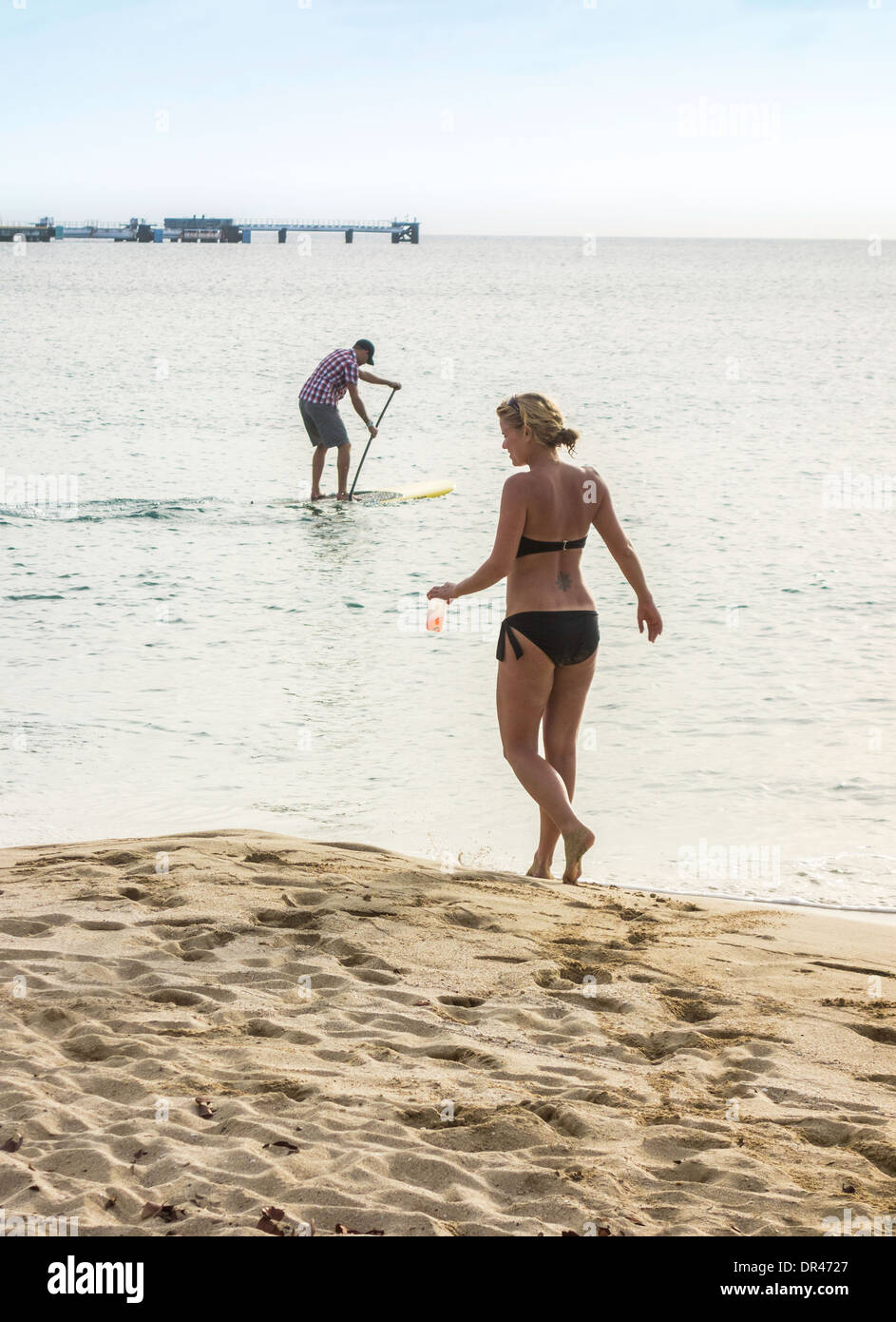 A Caucasian woman carries a drink along the beach while a man plays on a stand up paddle board in the Caribbean. St. Croix, U.S. Virgin Islands. Stock Photo
