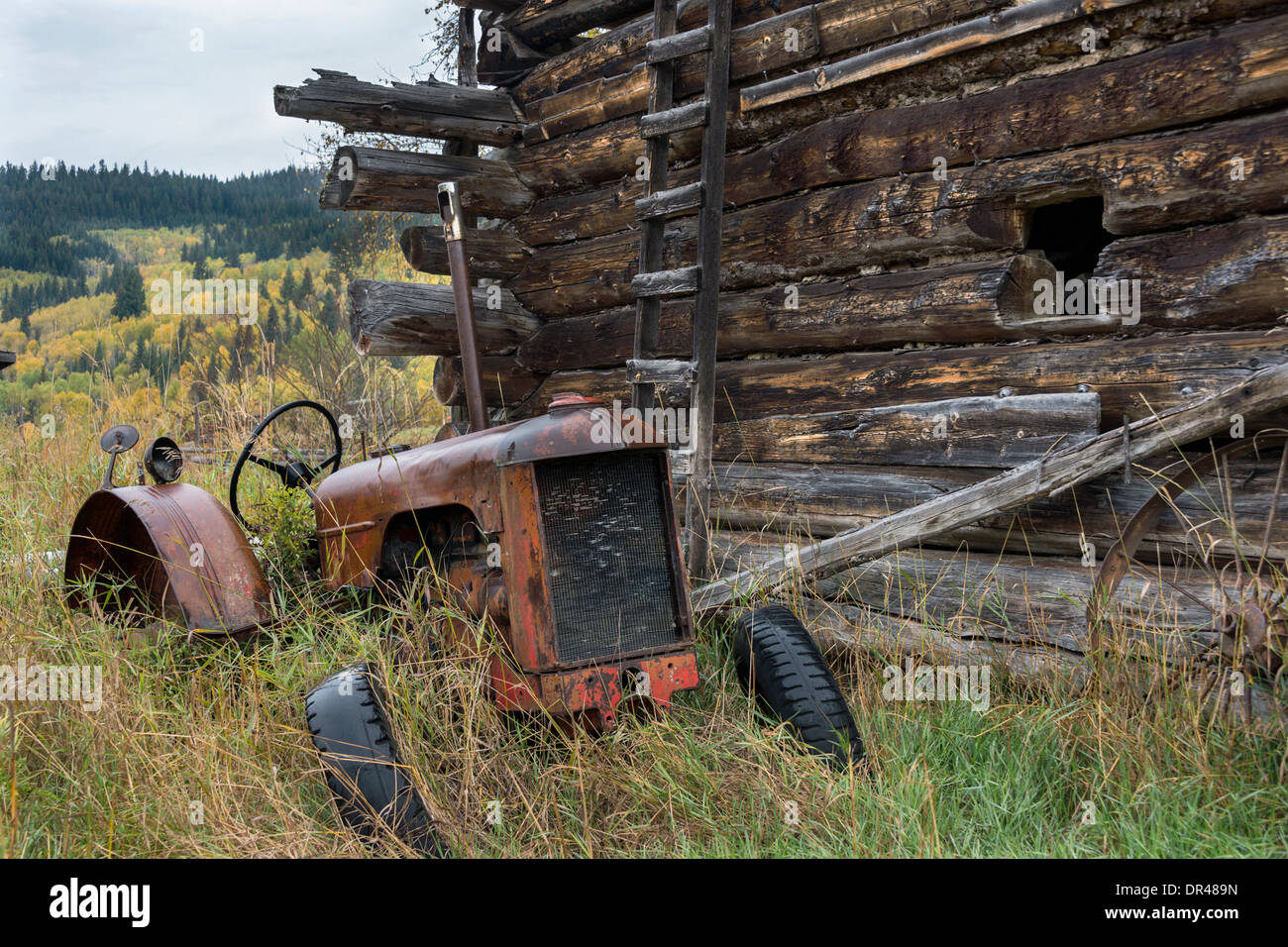 Old abandoned tractor by a log barn, on a farm near Likely, Cariboo-Chilcotin region, British Columbia Stock Photo