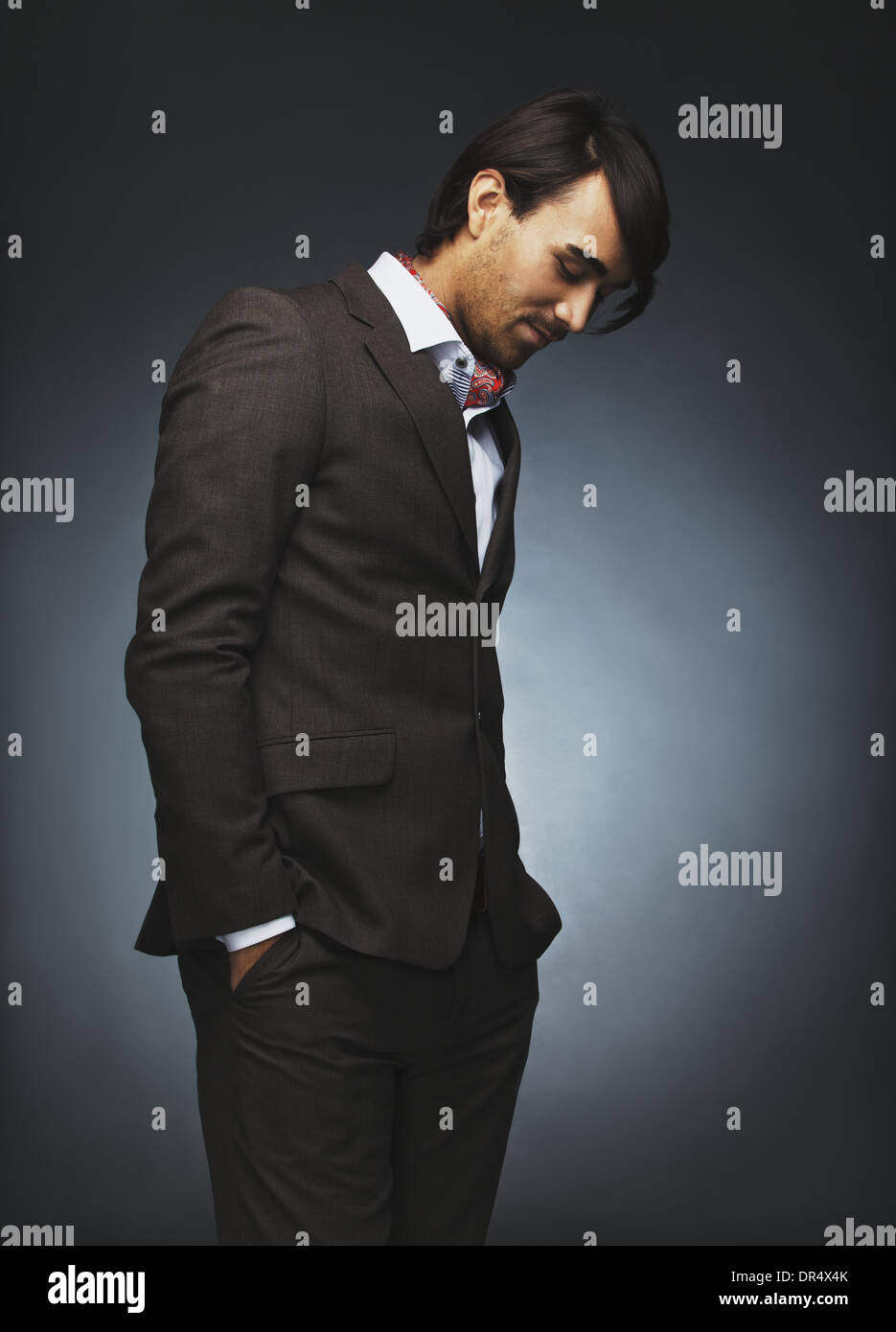 Attractive young man in stylish suit standing on black background. Male fashion model with hands in pocket looking down Stock Photo