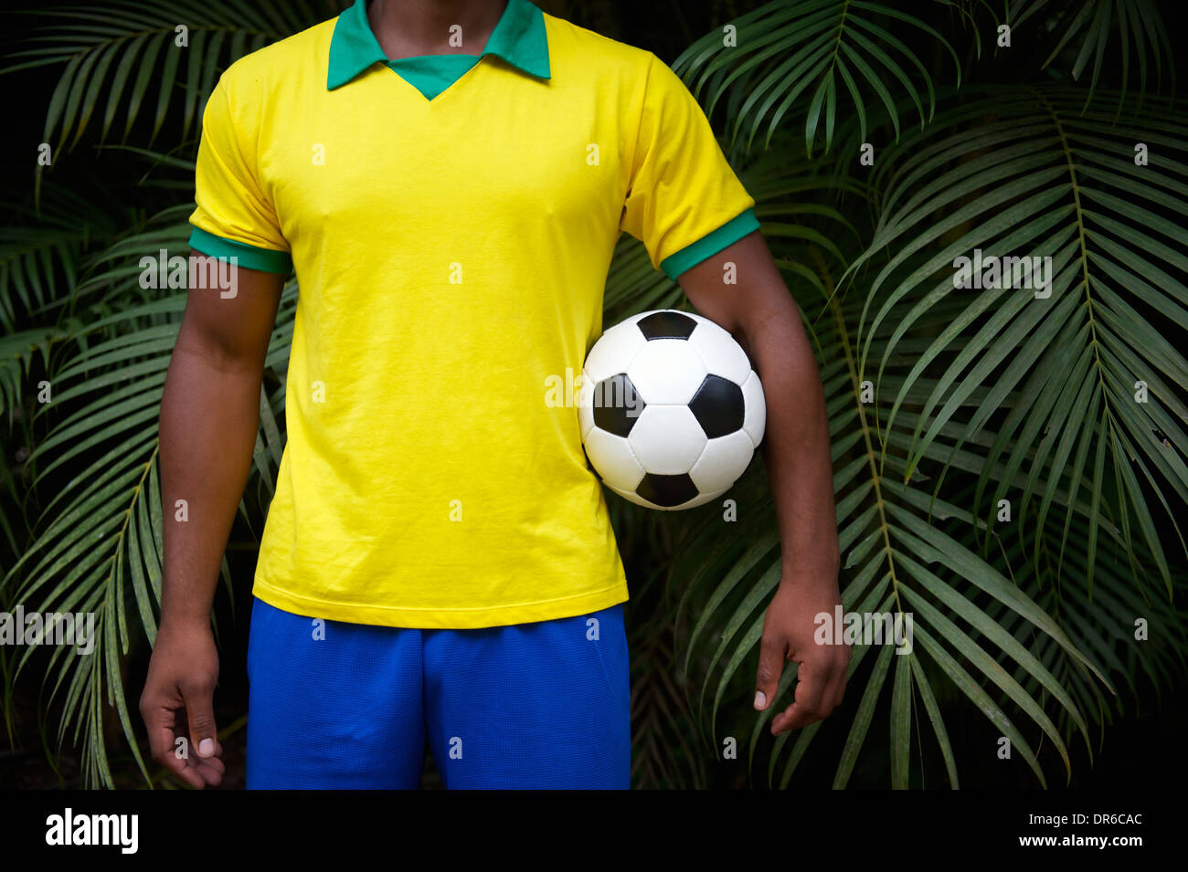Brazilian football player in Brazil colors uniform standing in tropical palm jungle holding soccer ball Stock Photo