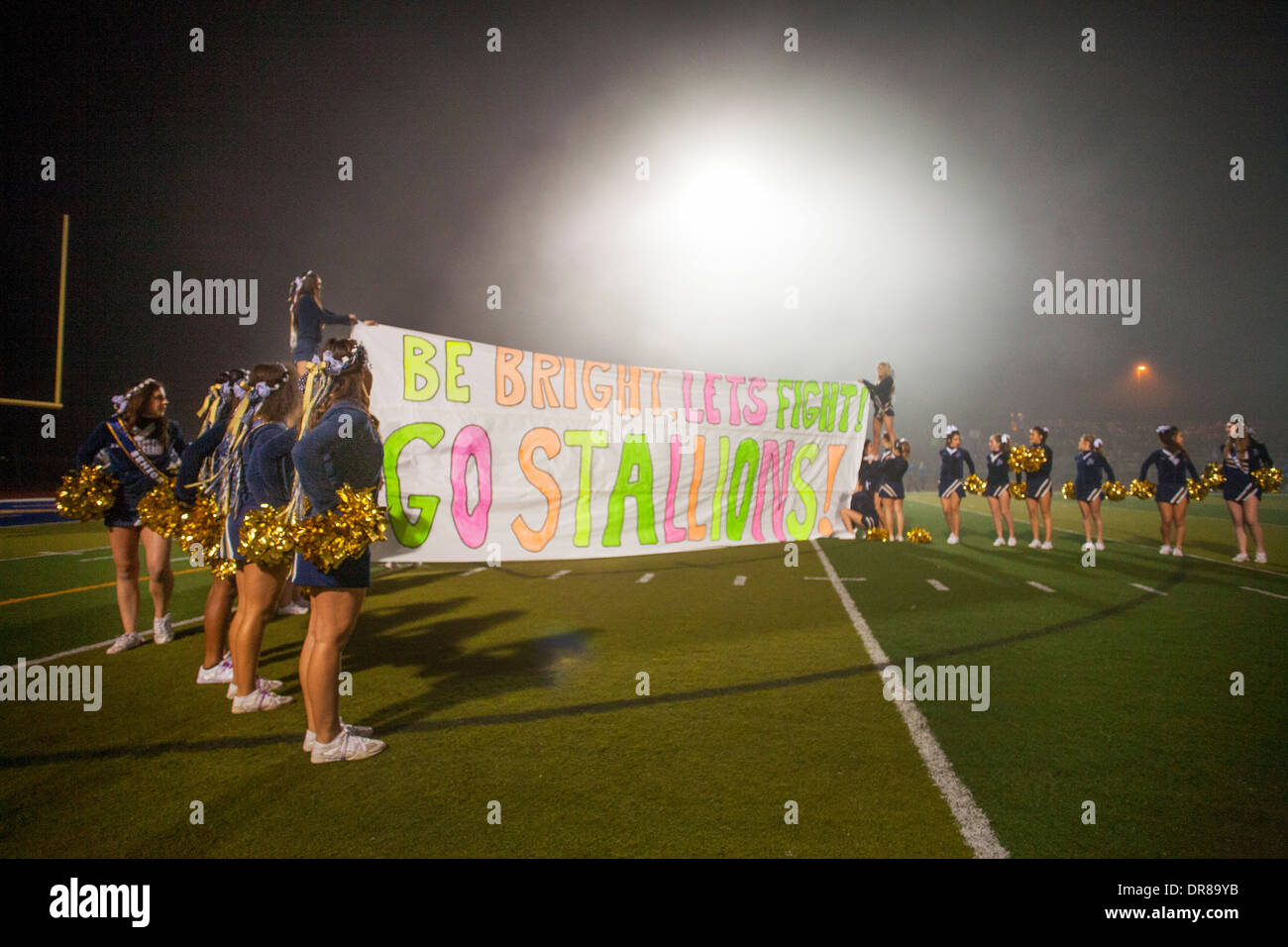 High school cheerleaders raise an inspirational banner to their football team before a night game in San Juan Capistrano, CA. Stock Photo