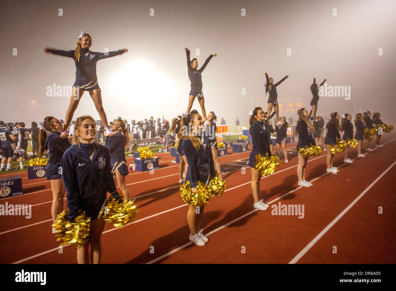 Cheerleaders entertain the spectators at a high school night football game in San Juan Capistrano, CA, hold initials of the school name. Note 'flyers' on shoulders of other cheerleaders. Stock Photo