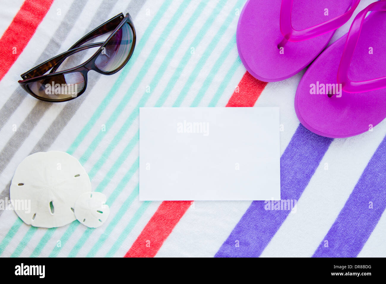 Beach scene with purple flip flops, sand dollars and sunglasses on a striped beach towel with copy space. Stock Photo