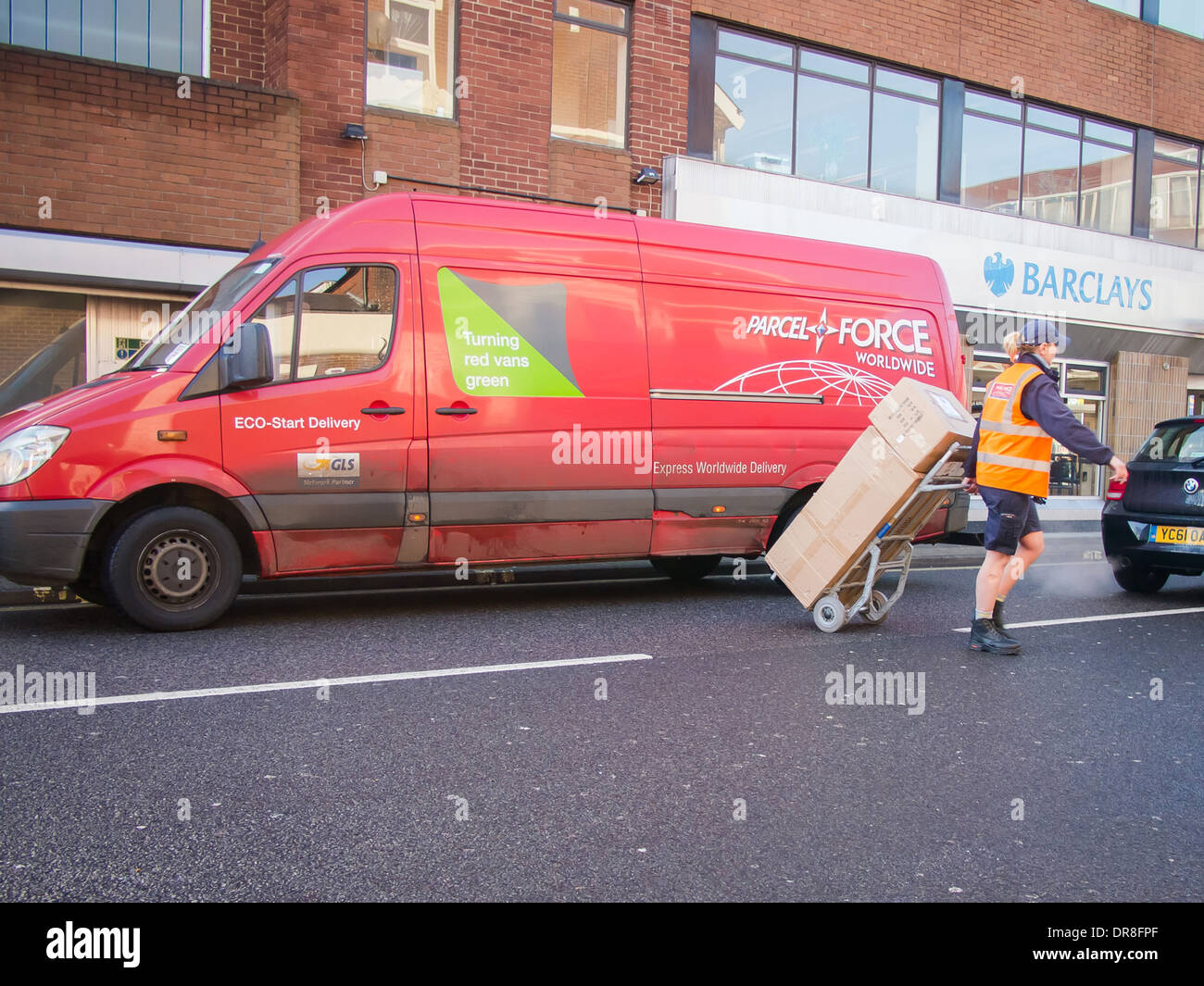 Parcel Force delivery van with delivery driver pulling stacked parcels across a road using a handtruck box carrier Stock Photo