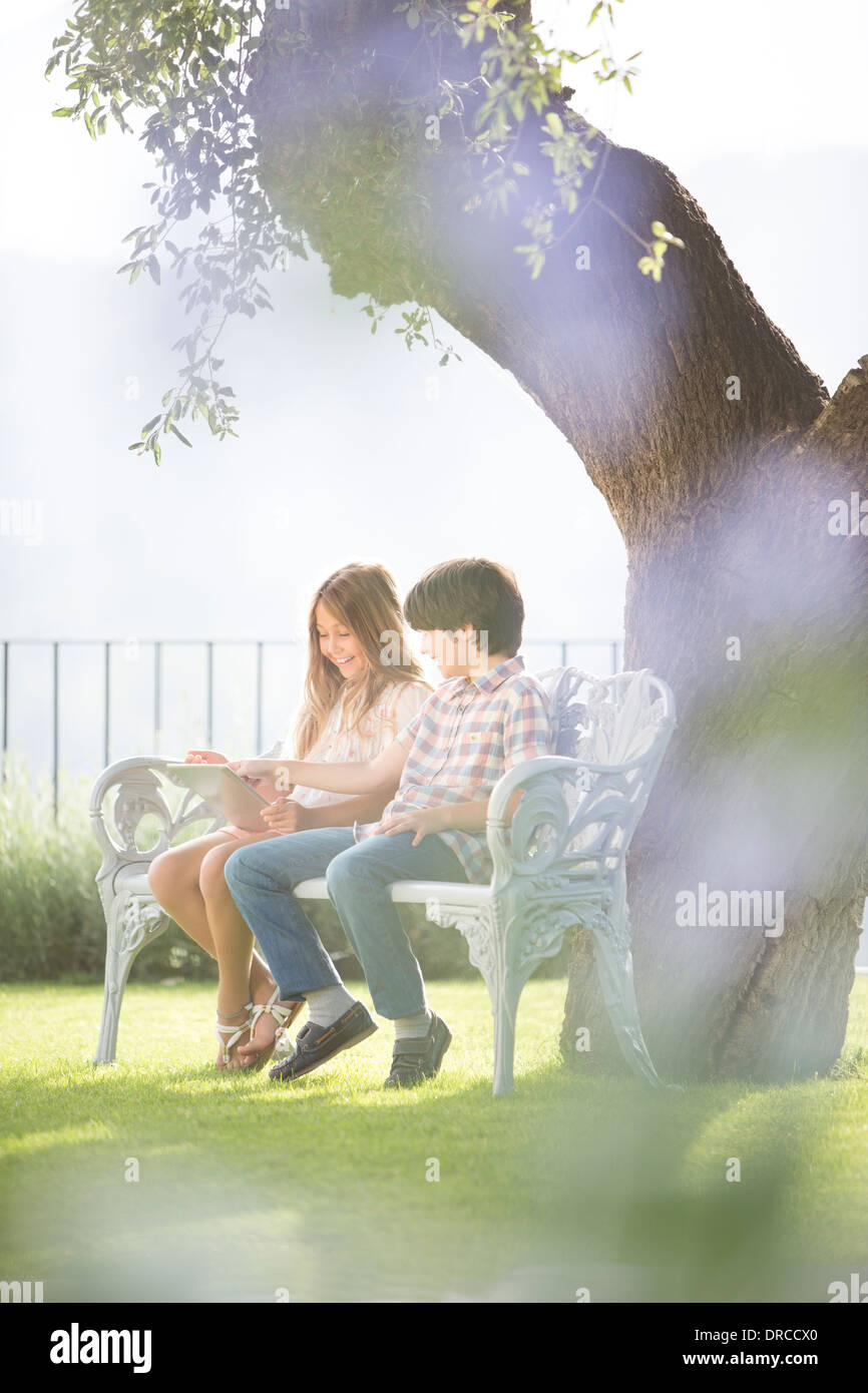 Brother and sister using digital tablet on bench under tree Stock Photo ...