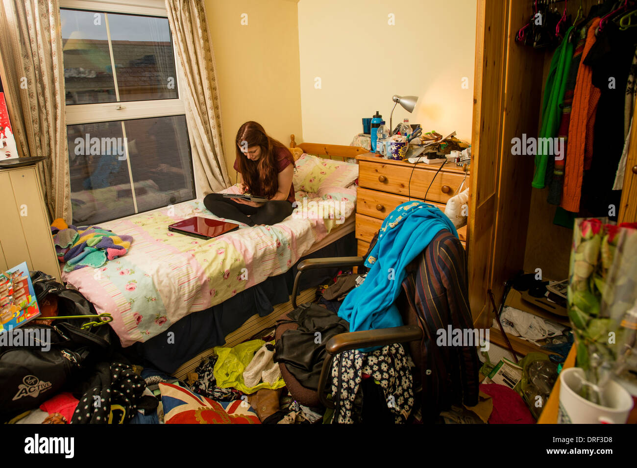 A young woman university student studying working on her iPad in her untidy bedroom at home UK Stock Photo