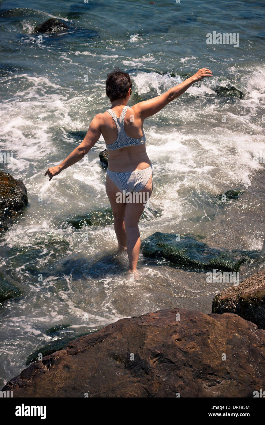 Middle aged woman walking in water on the rocky beach. Stock Photo