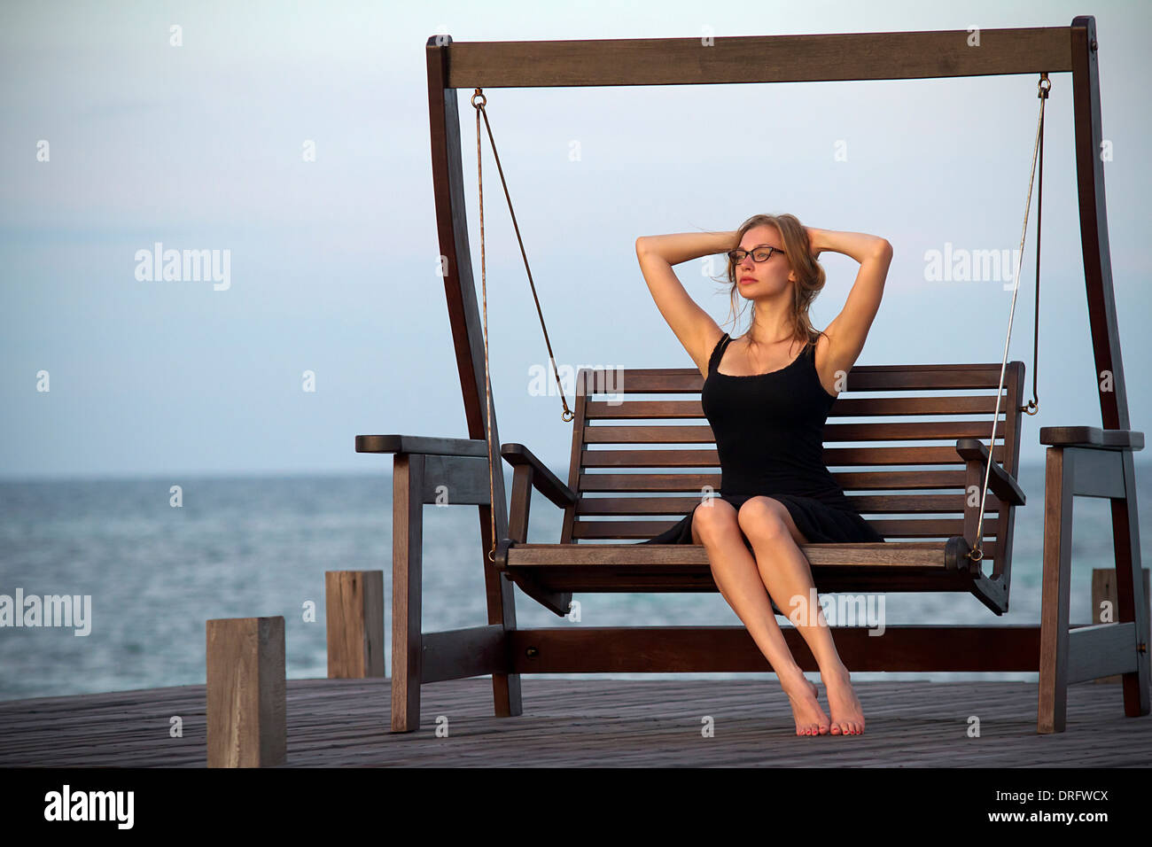 Young woman in sundress relaxes on a swing, hands behind head, Lankayan Island, Borneo, Malaysia Stock Photo
