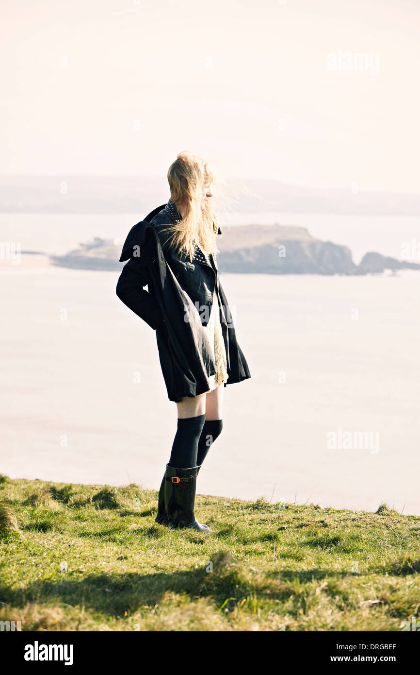 Fashion shoot of lady looking over water wearing long jacket, standing on cliff in Devon Stock Photo