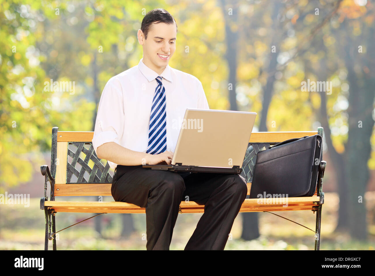 Young businessman sitting on a bench and working on a laptop in a park ...