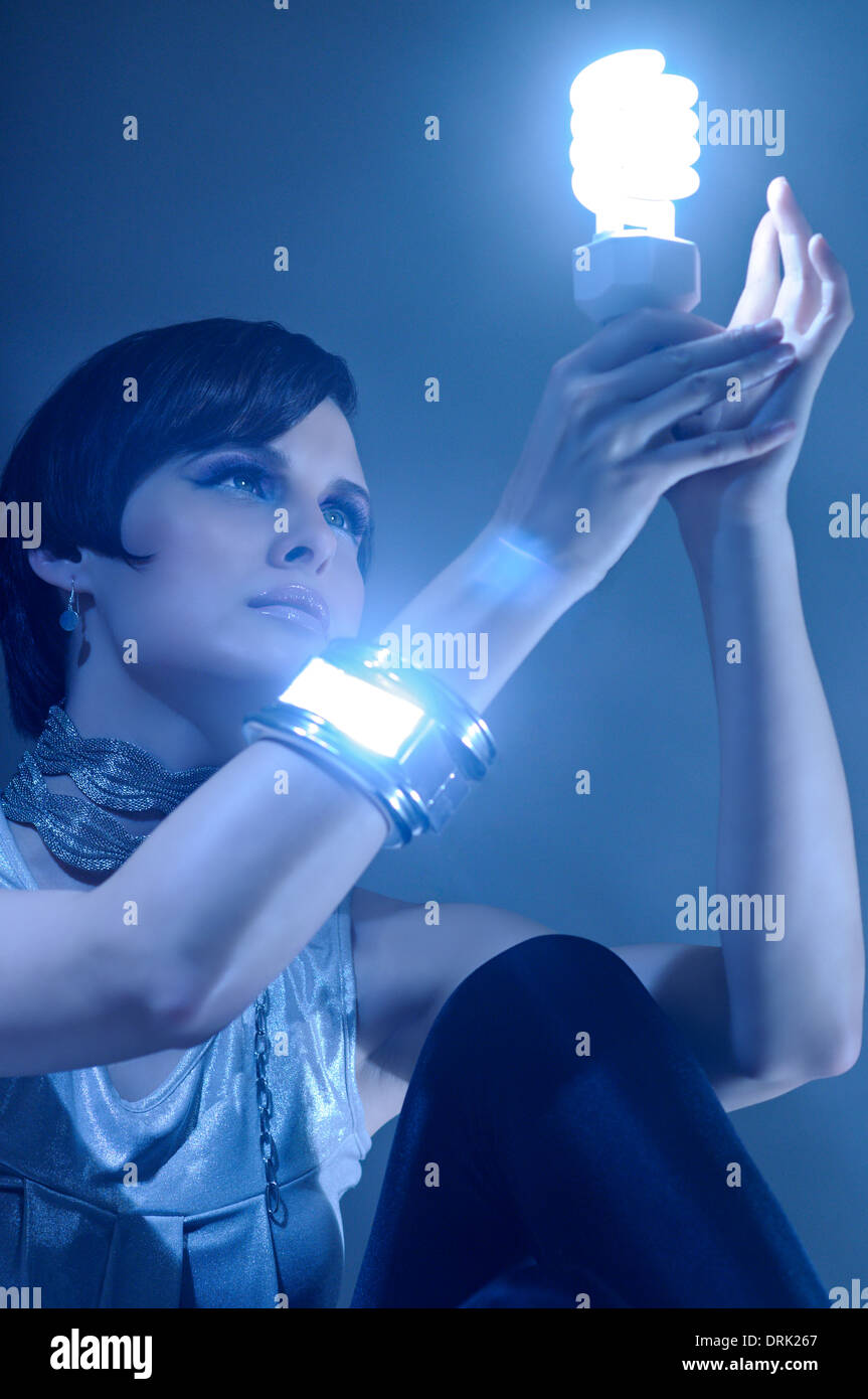 A beautiful futuristic woman holding a lit CFL bulb in her hands, looking at it Stock Photo