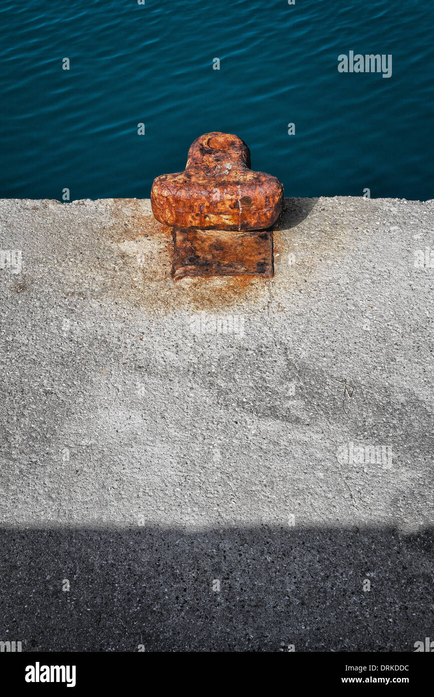 A semi-abstract image with a rust-encrusted bollard on a concrete quay. Band of rippled water at top, band of shadow at base. Stock Photo