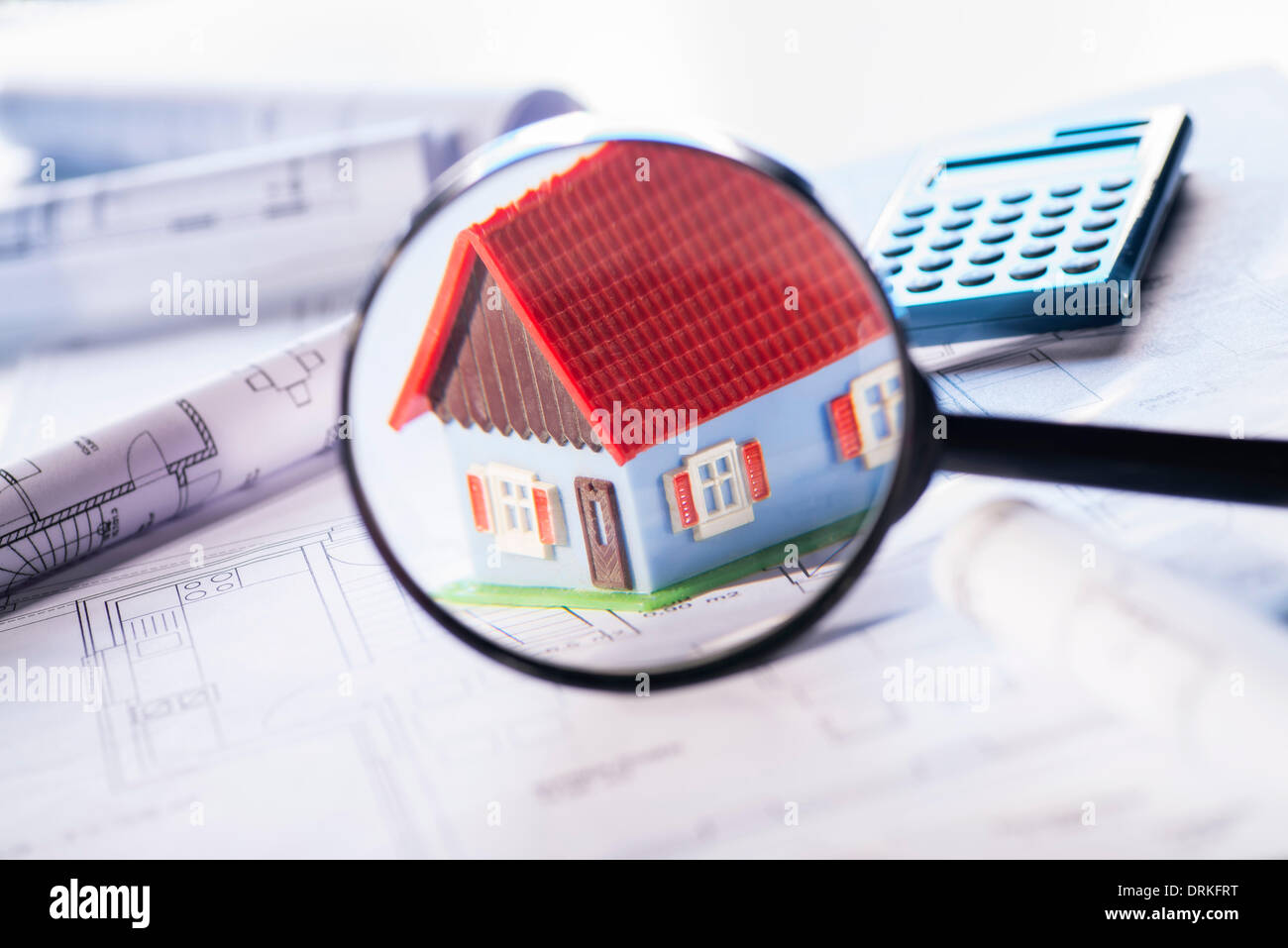 Architectural model is focused with a magnifying glass. In the background construction plans and a calculator can be seen. Stock Photo