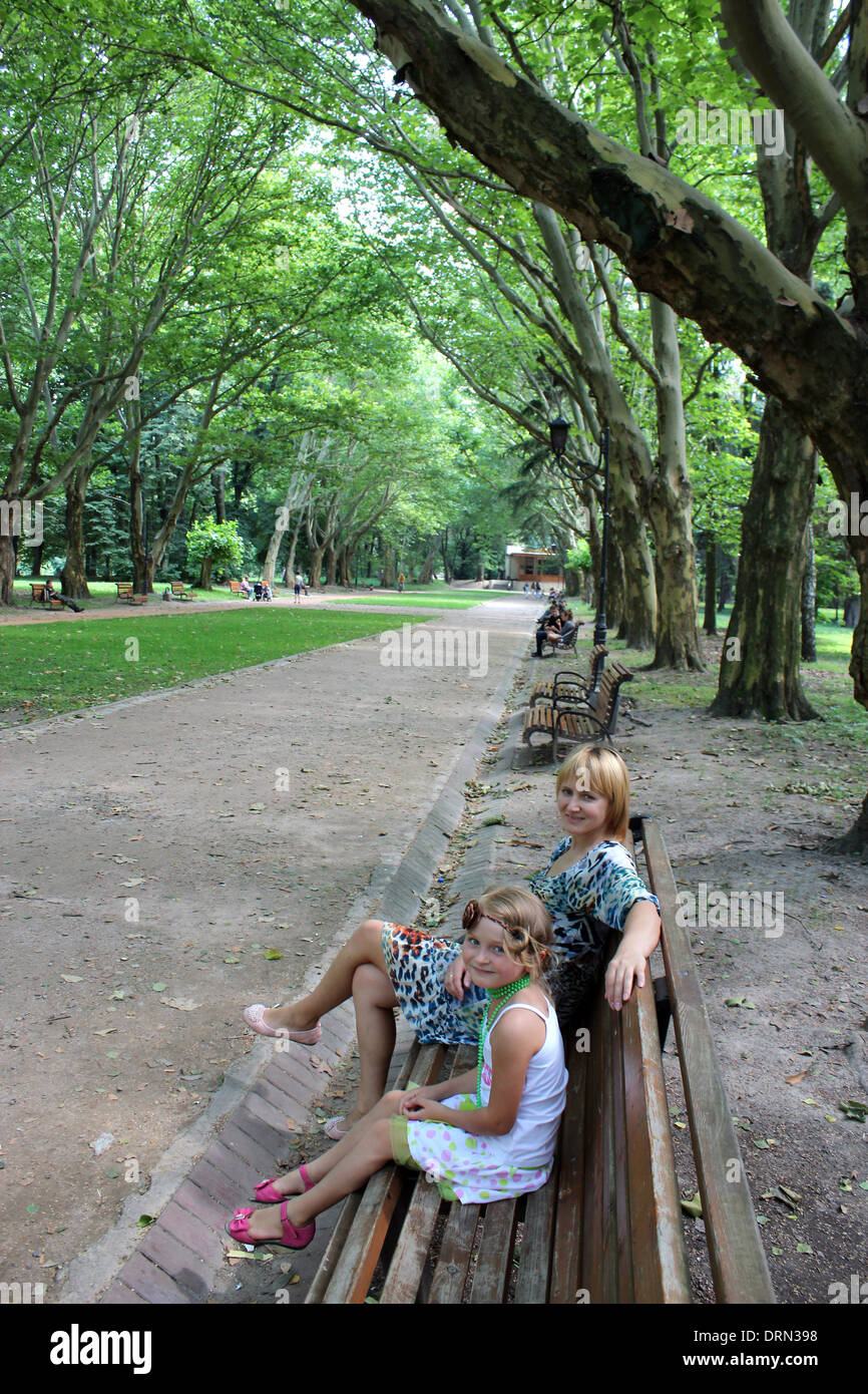 mother and daughter sitting on the bench in park with greater trees Stock Photo