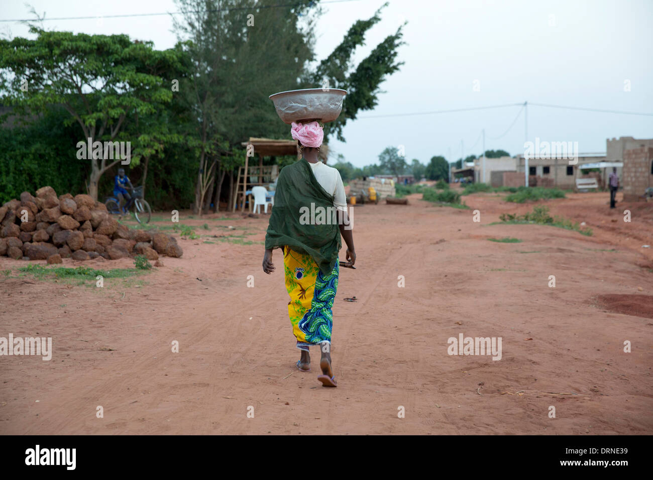 woman acrrying metal bowl on her head, walking down dirt road in Bobo Dioulasso, Burkina Faso, Africa Stock Photo