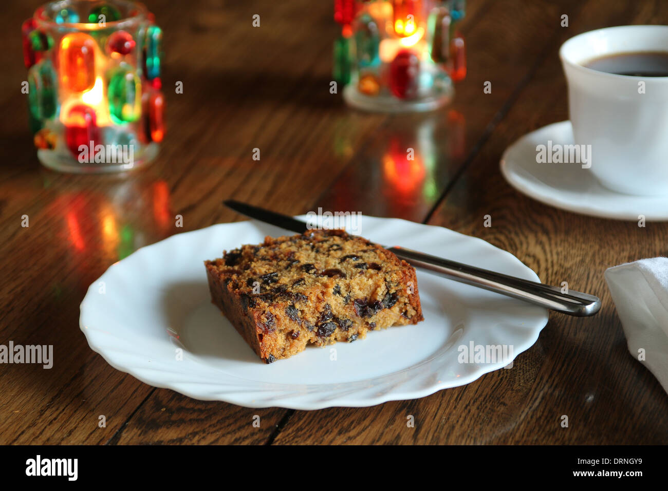 A slice of rich fruit cake, with no icing on a plate with a cup of coffee and candles in background (8 of a series of 8 Stock Photo
