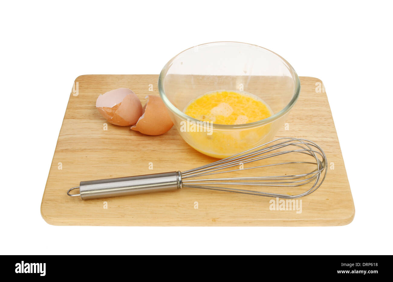 Egg in a glass mixing bowl with a whisk and egg shells on a wooden board isolated against white Stock Photo