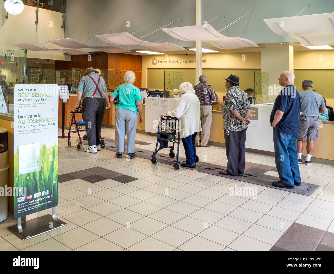 Elderly incoming patients line up to check in at the reception desk of a Southern California health maintenance organization. Note sign encouraging use of self service check in and senior man on walker. Stock Photo