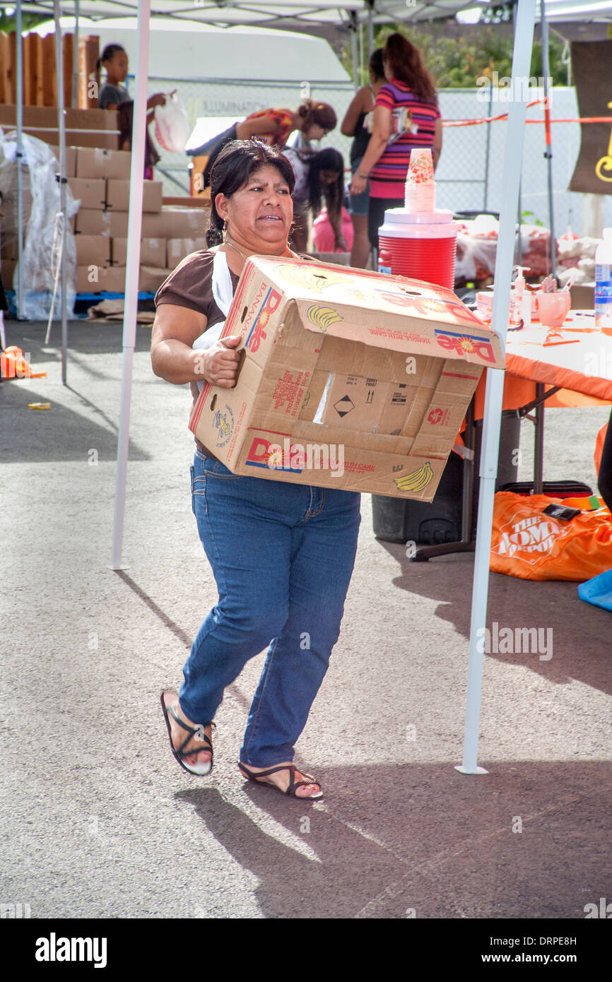 A hefty Hispanic housewife carries a heavy box of donated food from a charity site in a slum of Stanton, CA. Stock Photo