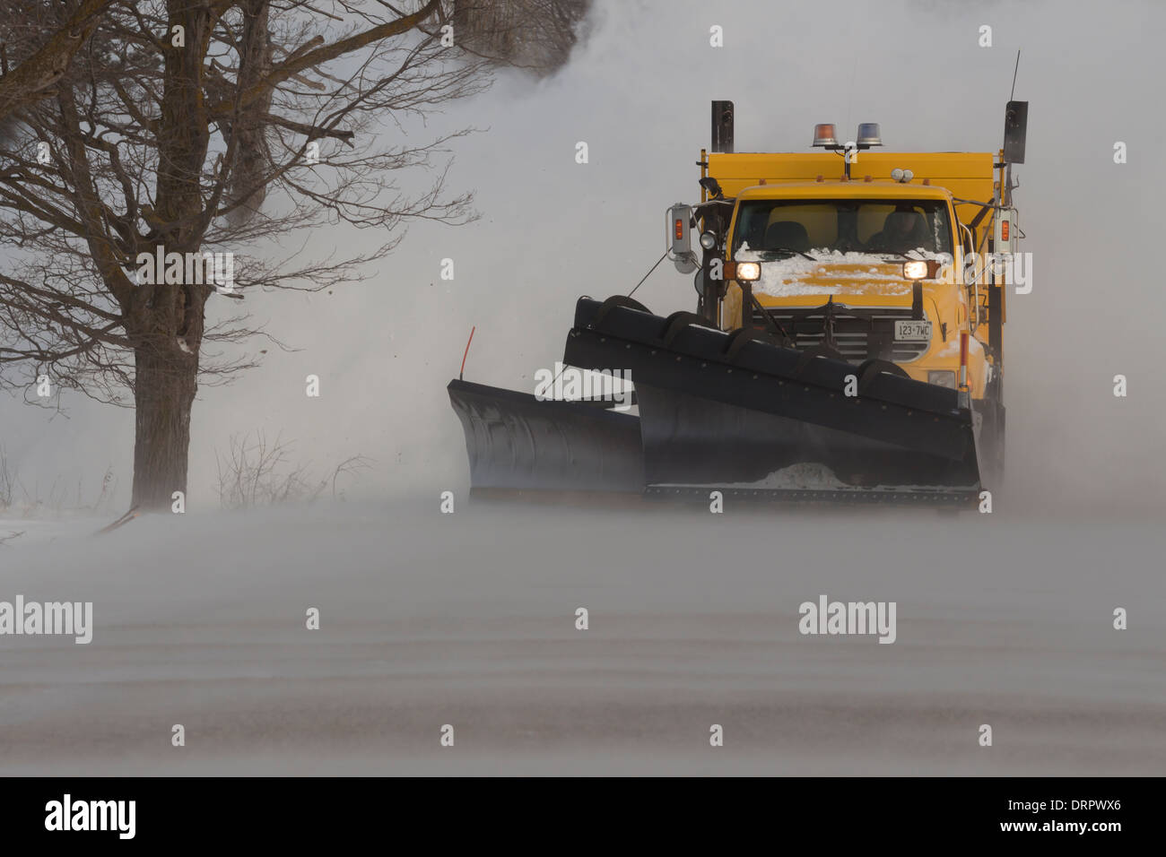 A yellow snow plow clears a road in blizzard conditions as wind blown snow covered the road creating large drifts Stock Photo