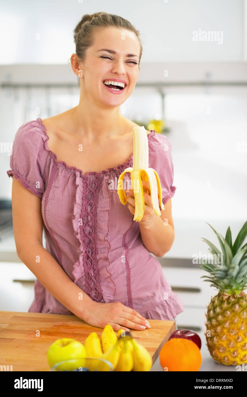 Smiling young woman eating banana in kitchen Stock Photo