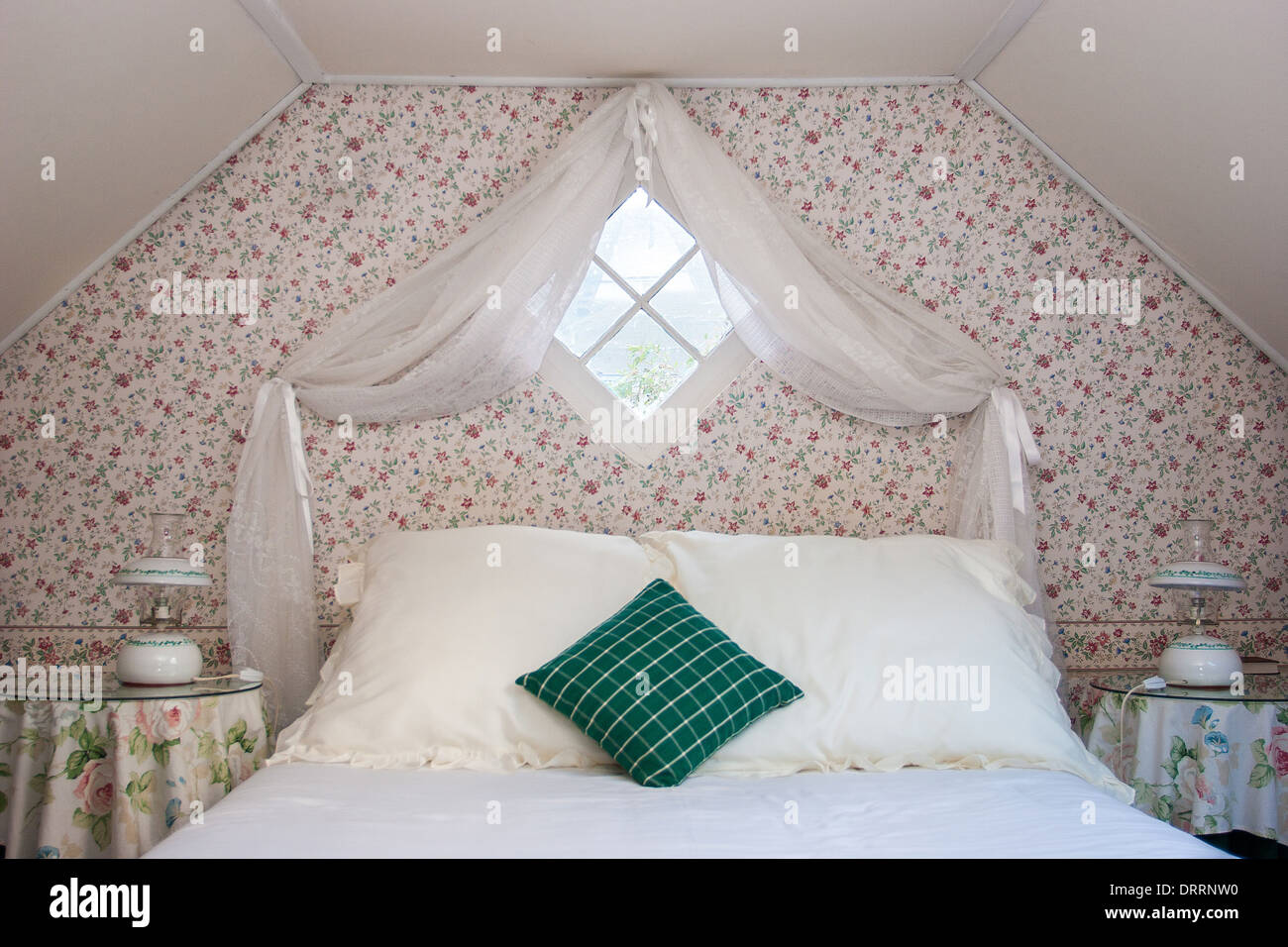 Symmetrical attic bedroom featuring curtained diamond window floral wallpaper and plain bed linen with emerald cushion Stock Photo