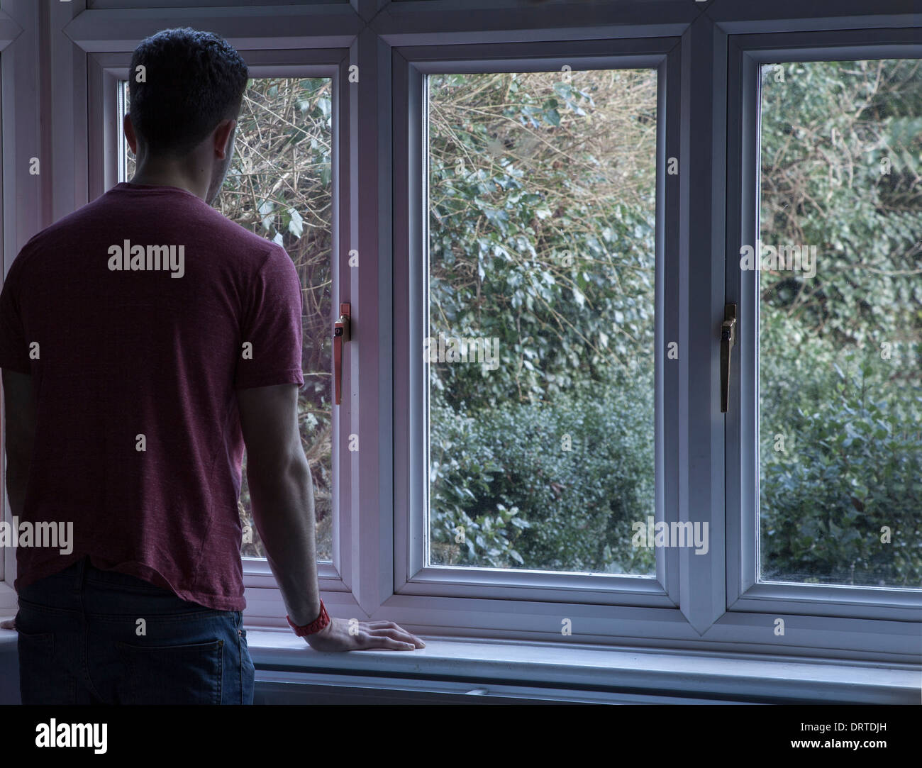 Young man indoors looking out through a window, over shoulder view. Stock Photo