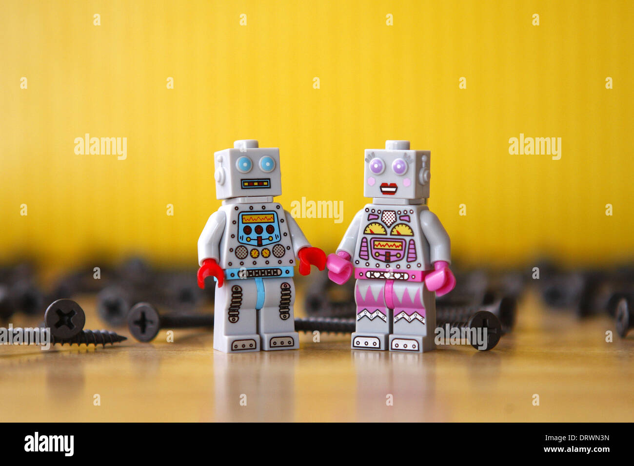 Grey male and female toy Lego robots surrounded by screws. Yellow background, wood floor Stock Photo