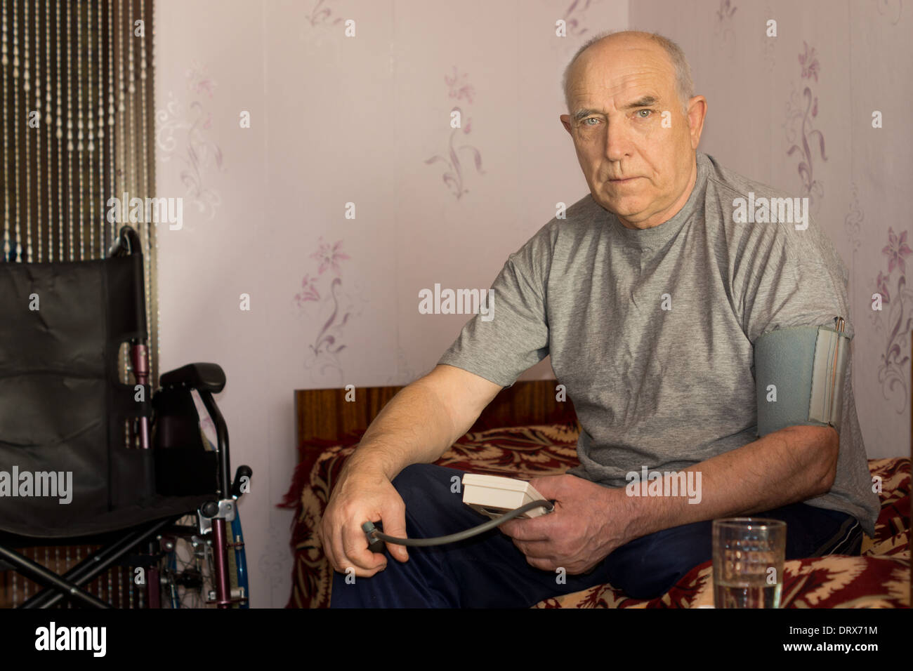 Senior man moniotoring his blood pressure using a pressure cuff and sphygmomanometer sitting on his bead at home Stock Photo