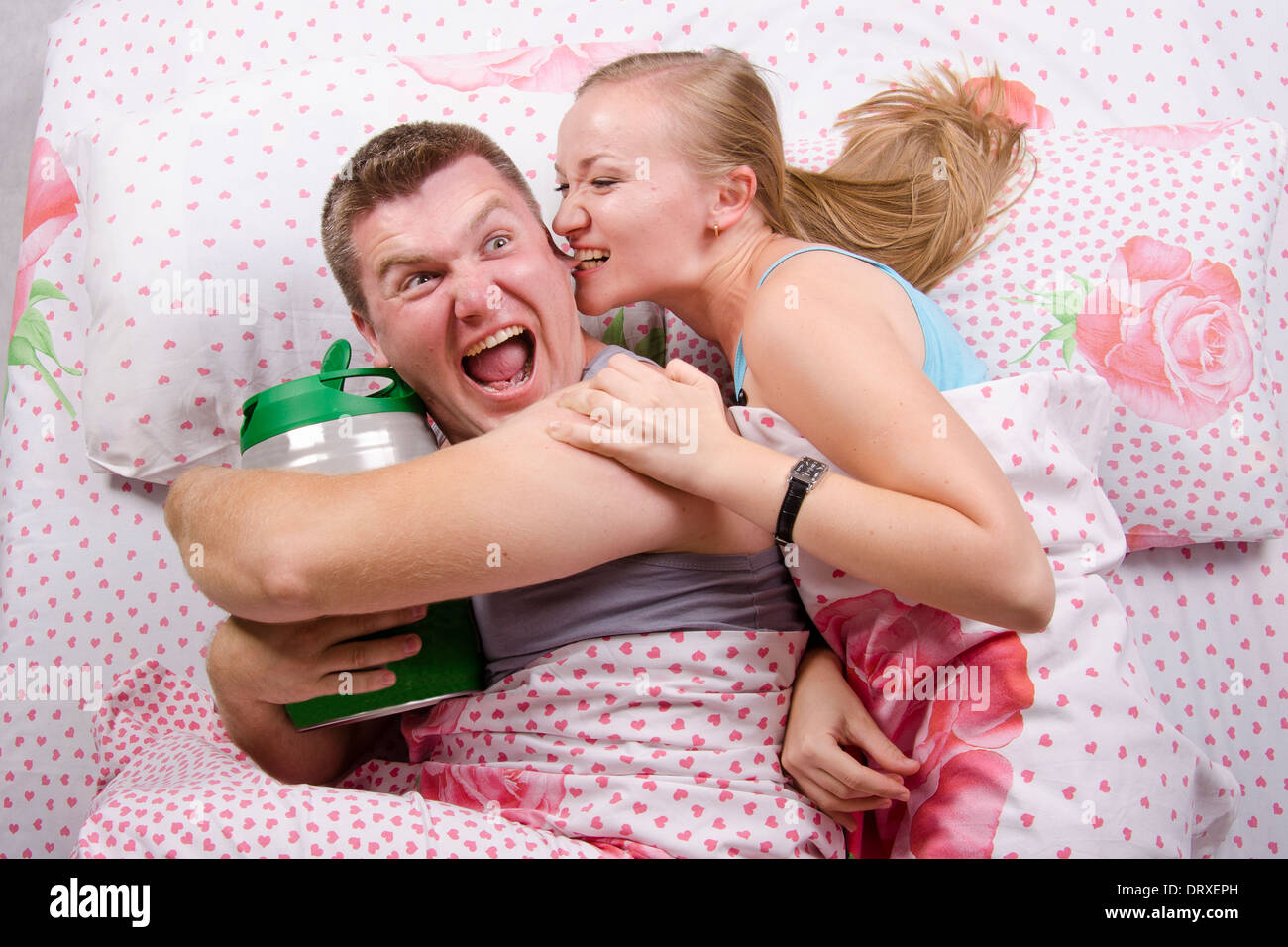Young couple lying in bed. Husband with a barrel of beer, wife trying to take barrel bites her husband's ear Stock Photo
