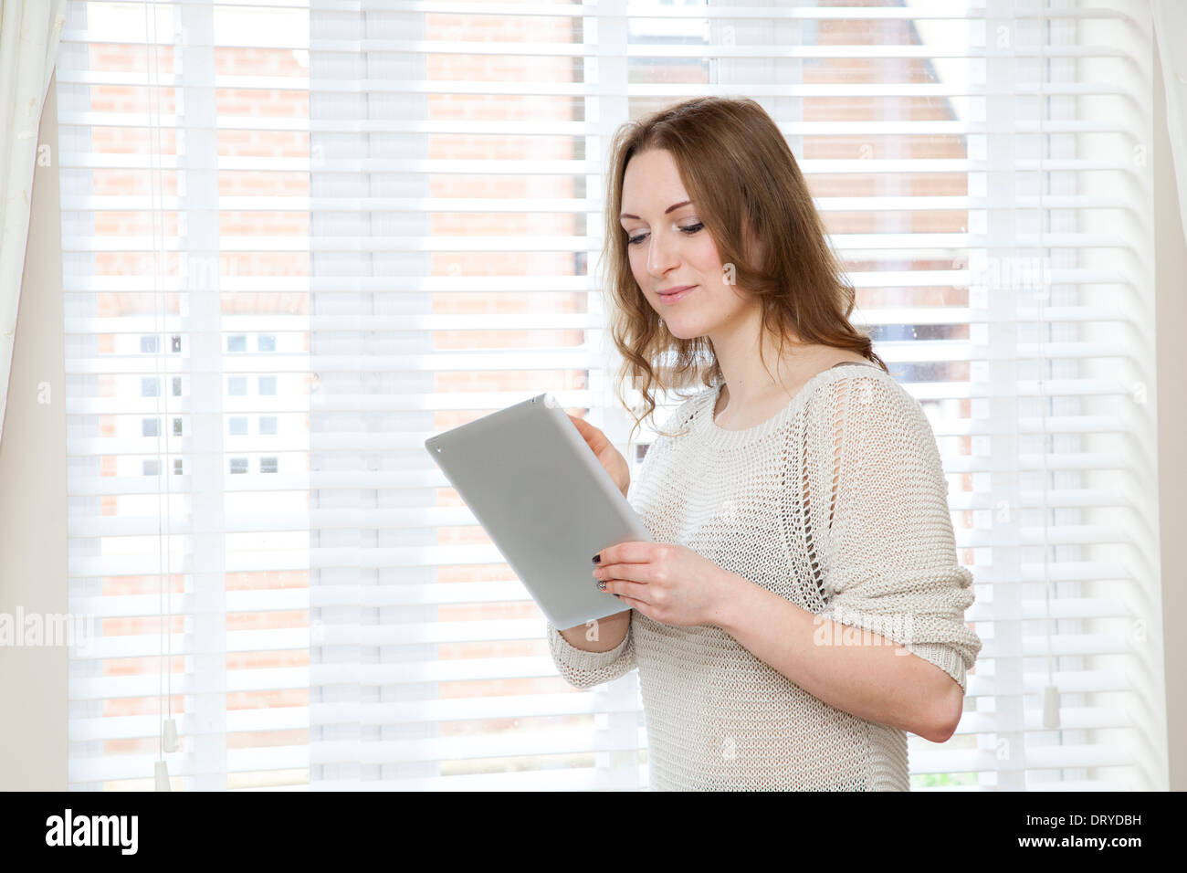 Young attractive woman holding a digital tablet (Ipad), by white window blinds. Stock Photo