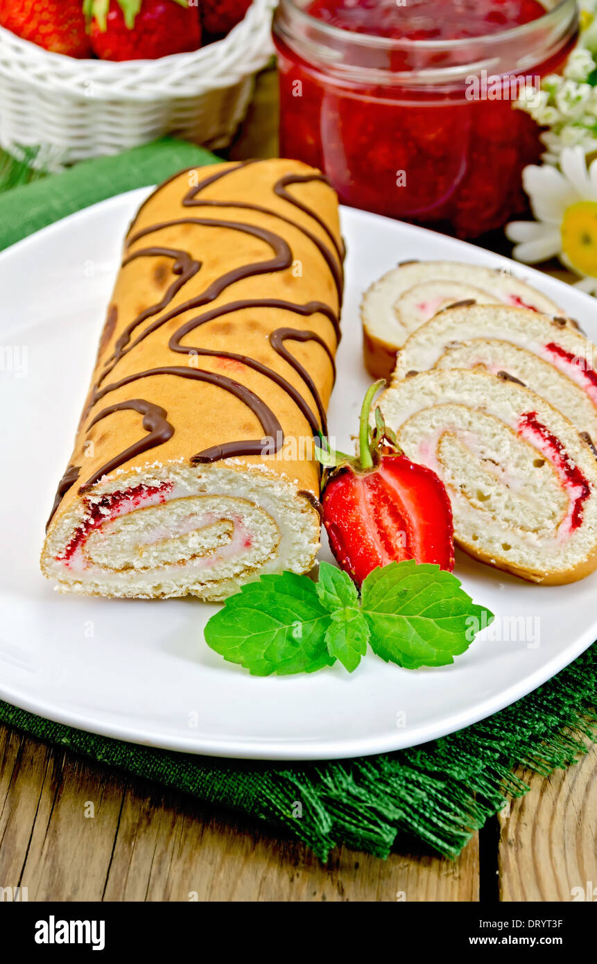 Biscuit roulade with cream and jam, jar of jam, doily, strawberries, mint on the background of wooden boards Stock Photo
