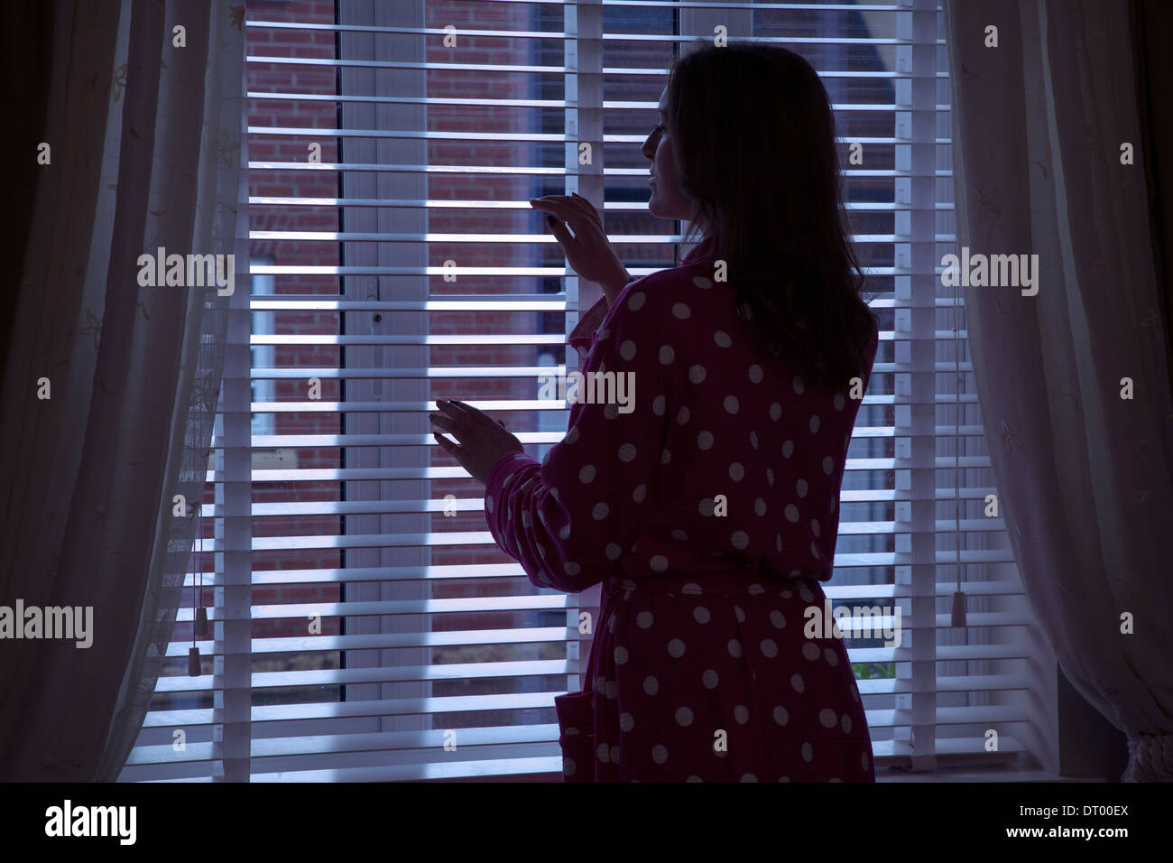 Silhouette of woman wearing a dressing gown looking out of a window at night. Over shoulder back/side view. Stock Photo