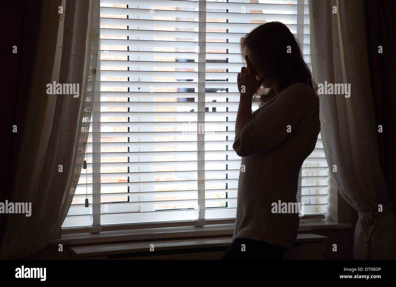 Silhouette of woman standing with her head in hands by a window. Over shoulder back/side view. Stock Photo