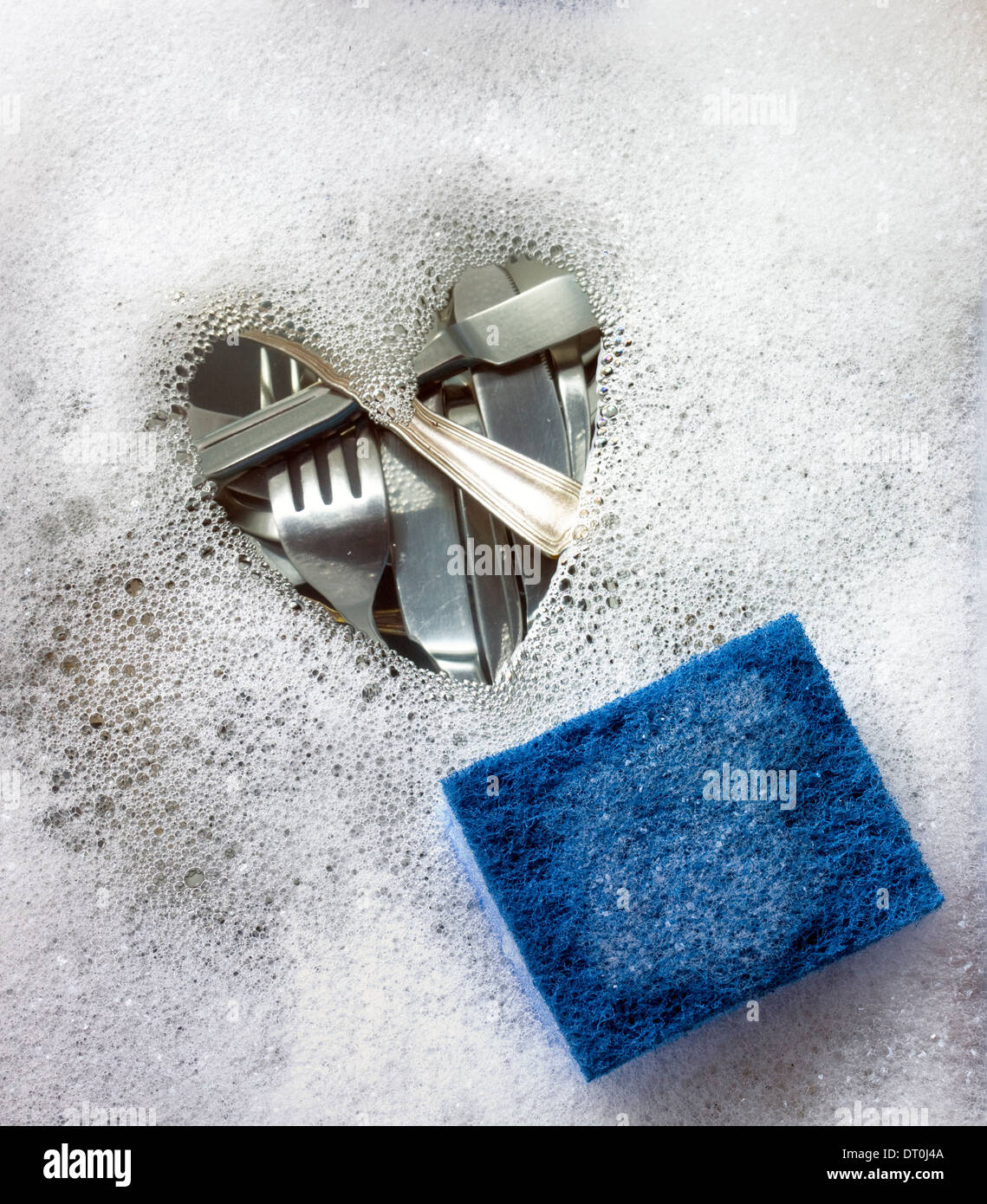 Heart shape in soap suds with cutlery beneath water and a blue scourer. Stock Photo
