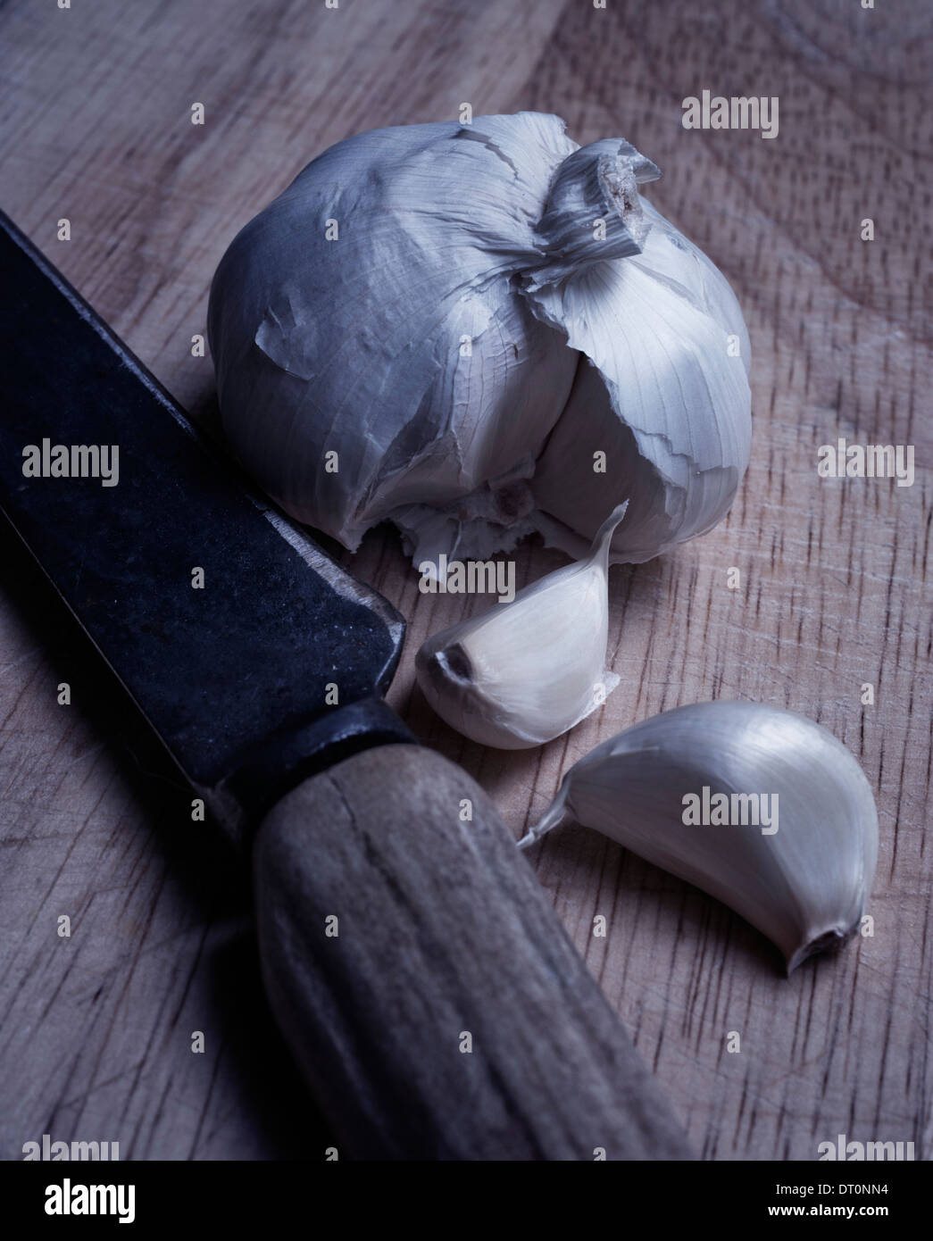 Garlic clove sits separately in front of garlic bulb on rustic wooden table. Rustic, bone-handled knife accompanies garlic. Stock Photo