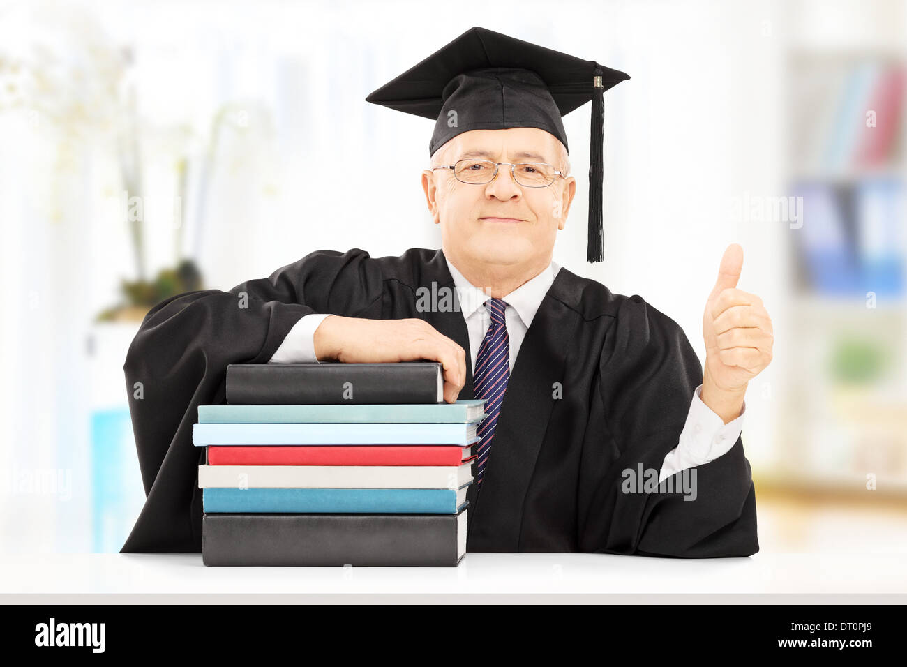 Middle aged college professor seated on table with stack of books and giving thumb up Stock Photo