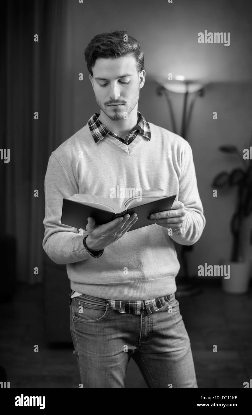 Handsome young man reading book at home in his living-room, standing. Black and white shot Stock Photo