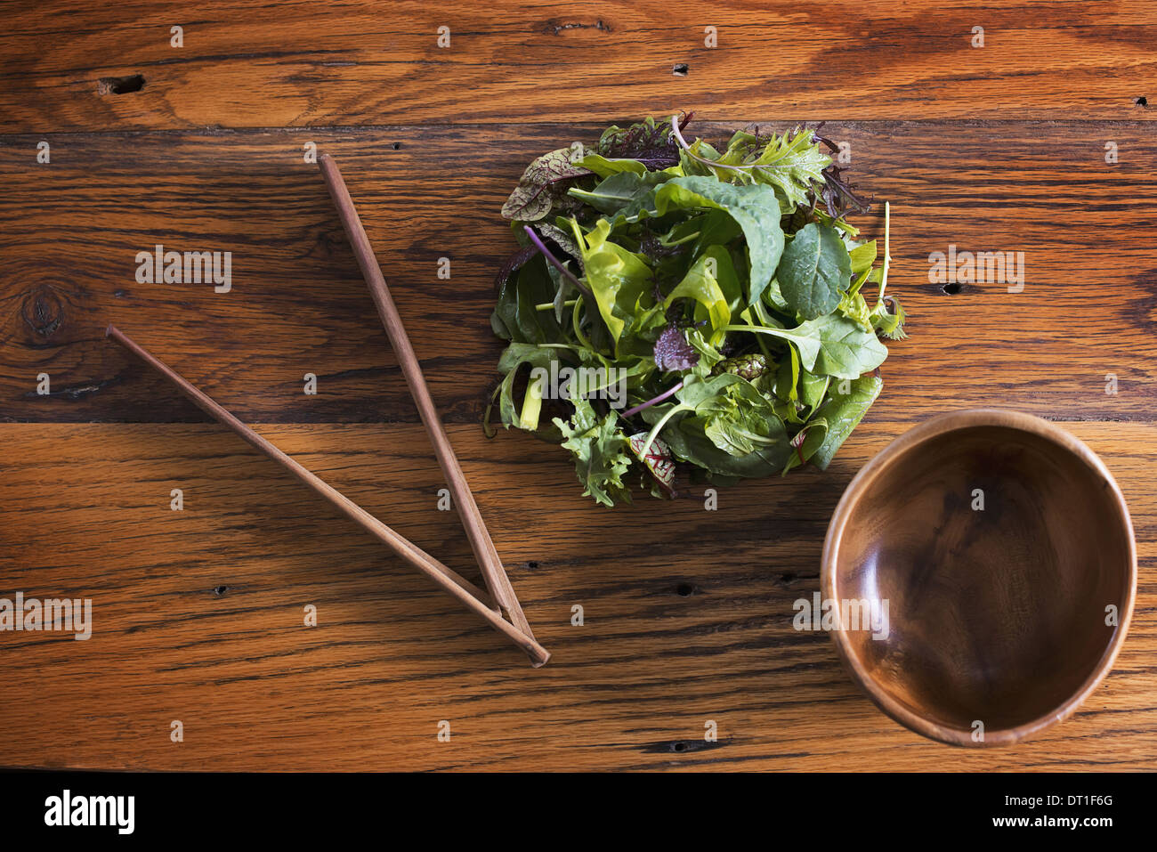A small round polished wooden bowl and a clutch of organic mixed salad leaves with wooden chopsticks Stock Photo