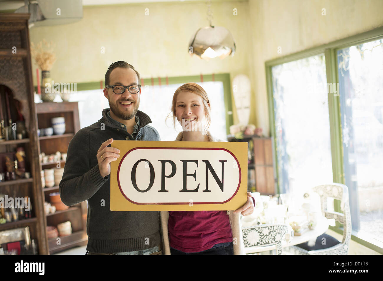 Two people standing in a store full of antique objects a couple running a business Holding a large sign saying OPEN Stock Photo