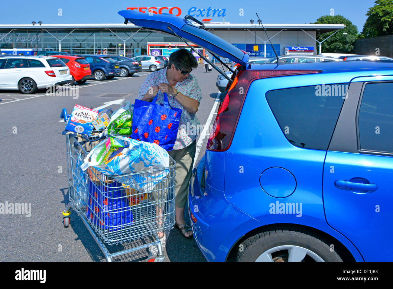 Tesco Extra supermarket store free car park mature woman load food shopping baskets from trolley into open hatchback car tail gate London England UK Stock Photo