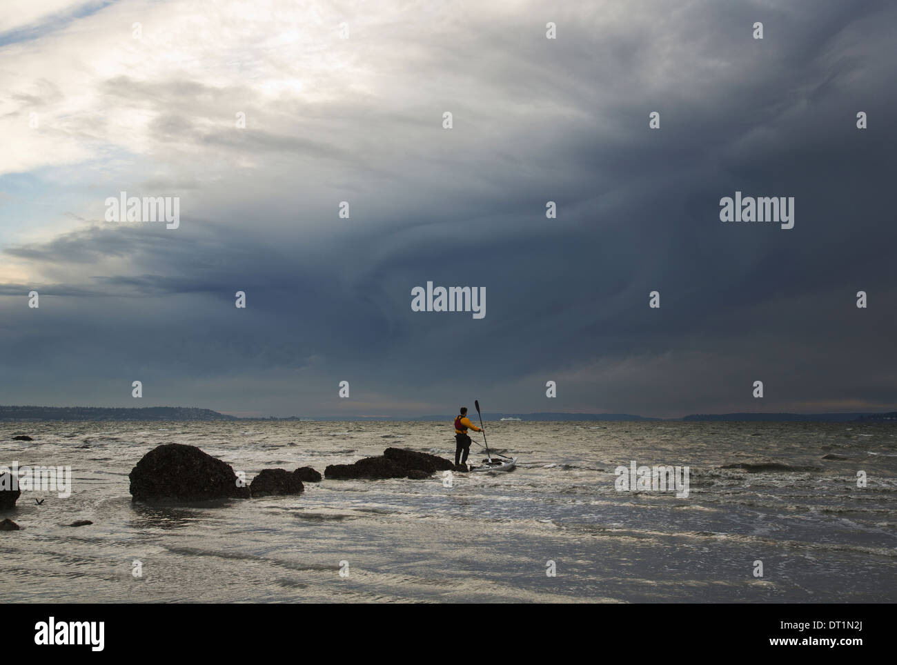 A man stands on a rock in the middle of the Puget Sound with his sea kayak floating next to him Stock Photo