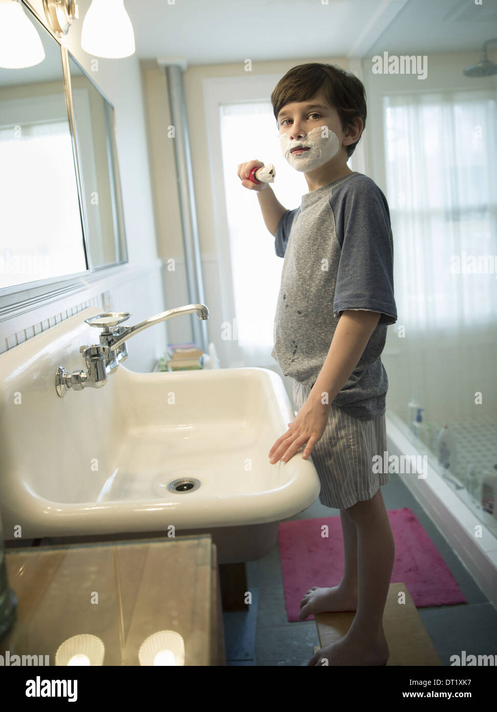 A young boy standing in a bathroom holding a shaving brush covering his face with foam Stock Photo