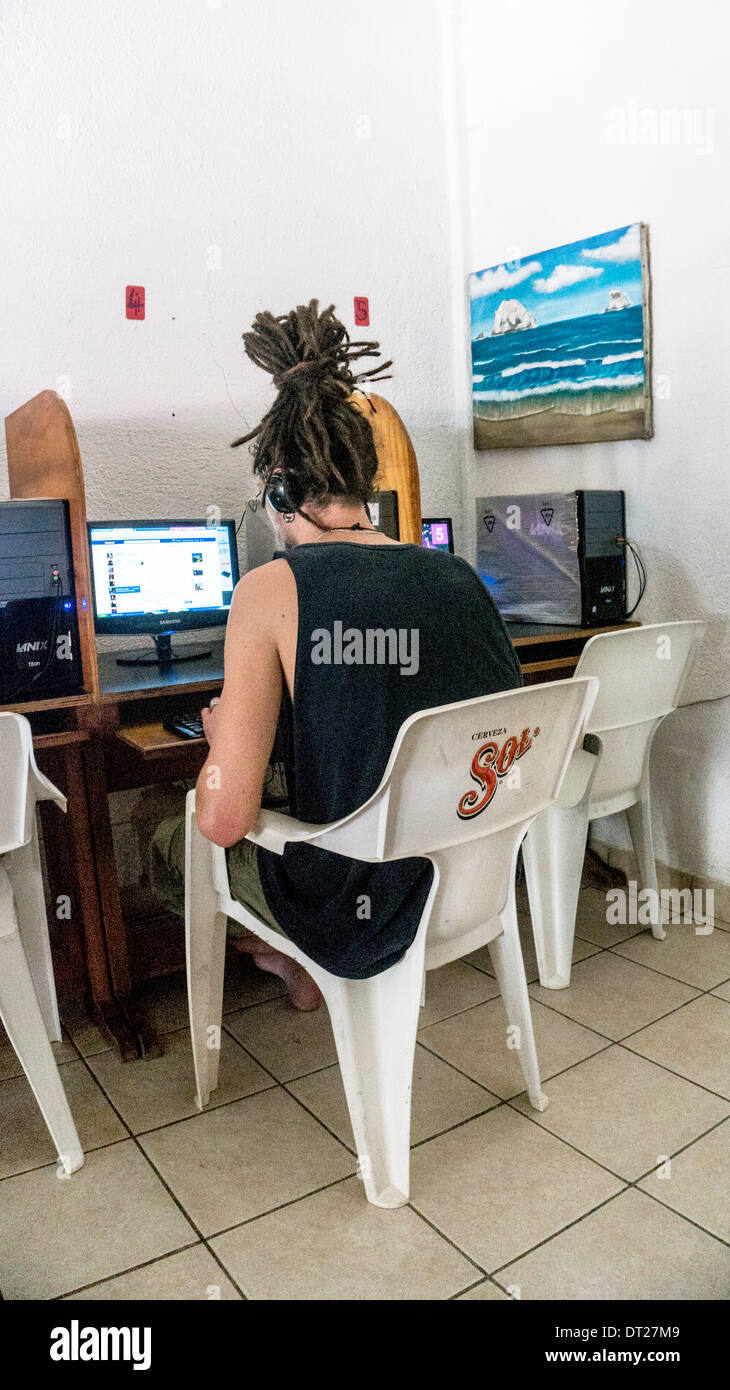 young man with dreadlocks piled atop his head sits at public access computer looking at Facebook in internet cafe Zipolite Stock Photo