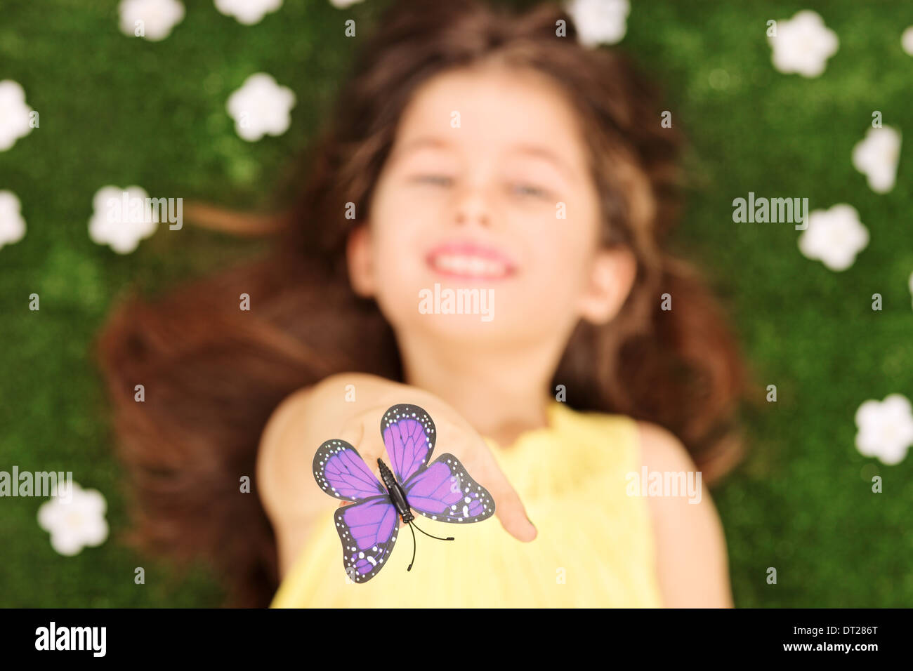 Blurred photo of little girl laying in meadow with flowers and reaching her hand to touch a purple butterfly Stock Photo