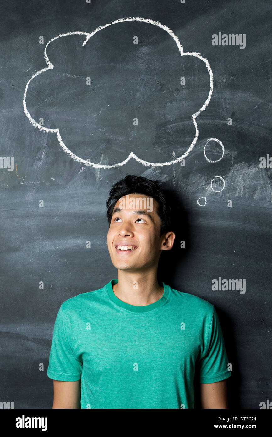 Happy Asian man standing under thought bubble hand drawn on a dark chalkboard. Stock Photo