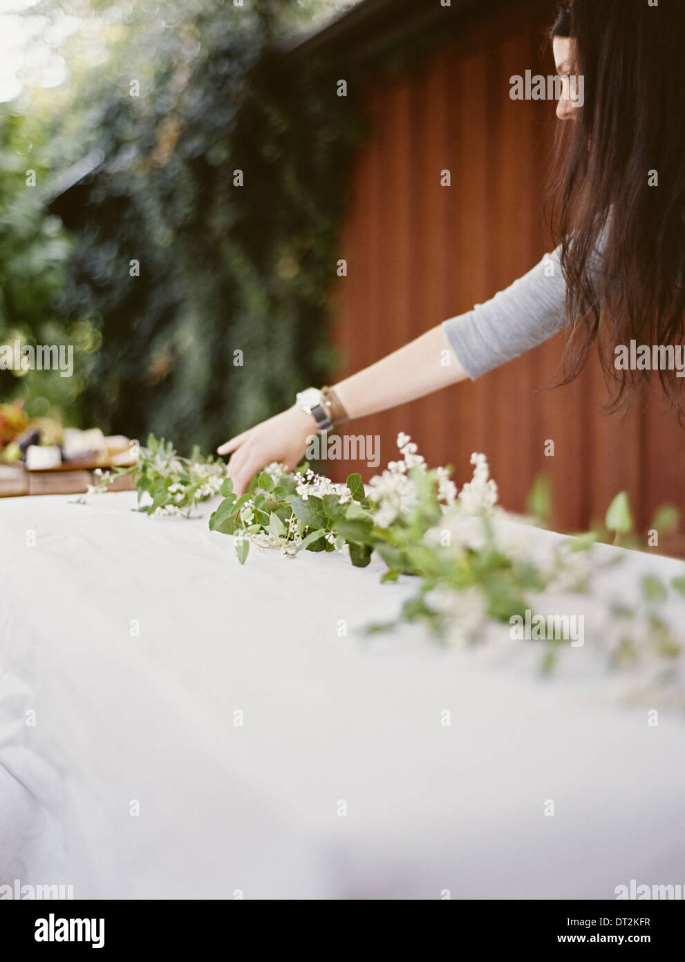 A woman with long hair by table laid outside with a white cloth and central foliage table decoration Place settings Stock Photo