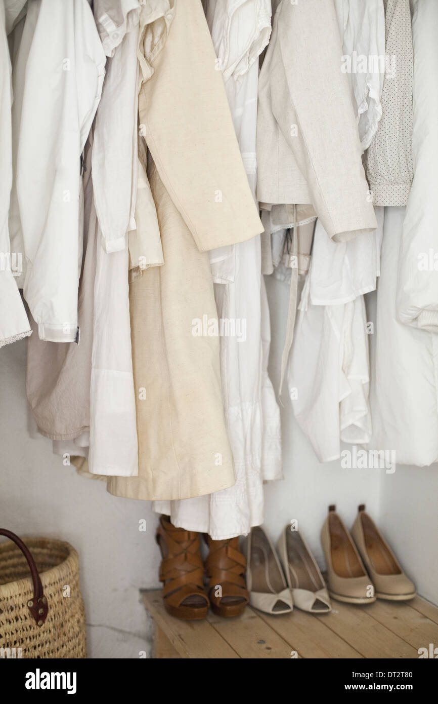 A closet with cream and white clothes jackets shirts and tunics hanging up A row of women's shoes arranged neatly on the floor Stock Photo