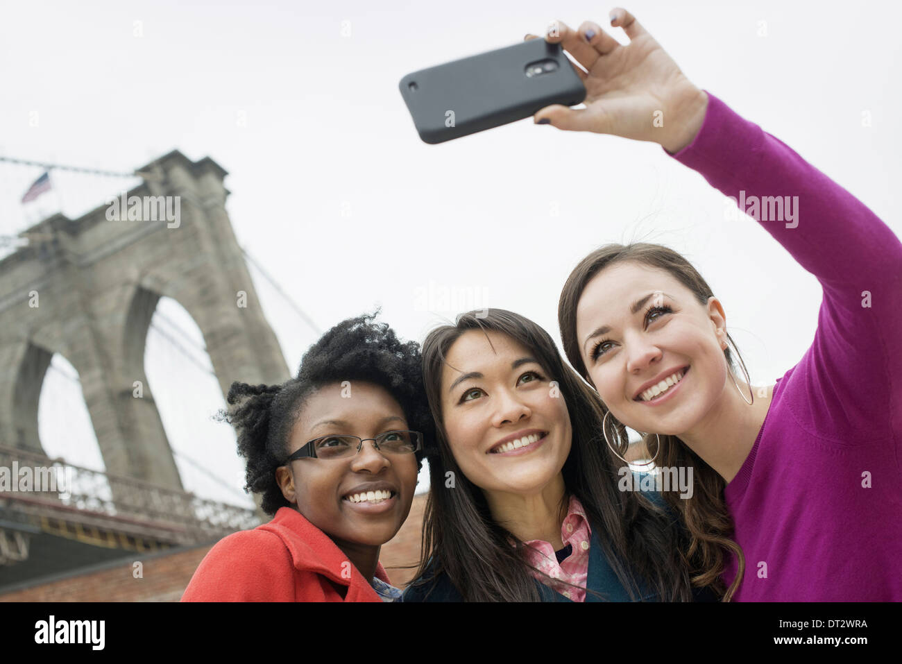 New York city the Brooklyn Bridge East River Three women in a row smiling as one takes a picture with a smart phone Stock Photo